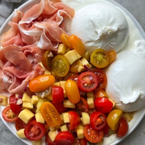 A plate with prosciutto slices, two whole balls of fresh mozzarella, and a colorful mix of halved cherry tomatoes and diced yellow vegetables makes for health-conscious bites inspired by classic food recipes.