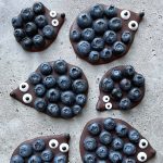 Six chocolate bites resembling hedgehogs are topped with blueberries and candy eyes, arranged on a gray textured surface. Each “hedgehog” features a pointed nose and clusters of blueberries as “spines”—a fun idea for health-focused food recipes.