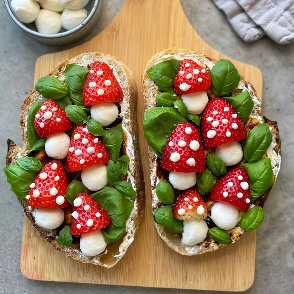 Two slices of rustic bread topped with strawberries, mozzarella balls, basil leaves, and cream dots create vibrant, fresh bites on a wooden board. A bowl of mozzarella sits nearby for extra flavor and inspiration for your healthy food recipes.