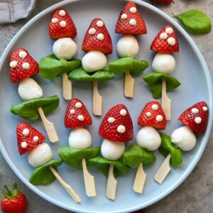 A plate of healthy snacks arranged to look like mushrooms, made from strawberries, mozzarella balls, basil leaves, and wooden sticks, with dots of cheese on the strawberries for decoration—perfect bites inspired by creative food recipes.