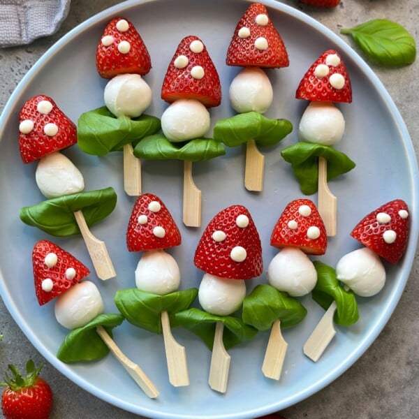 A plate of healthy snacks arranged to look like mushrooms, made from strawberries, mozzarella balls, basil leaves, and wooden sticks, with dots of cheese on the strawberries for decoration—perfect bites inspired by creative food recipes.