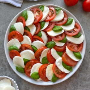 A plate of tomato caprese salad arranged in a circular pattern with alternating slices of fresh mozzarella, ripe tomatoes, and basil leaves on a gray surface.
