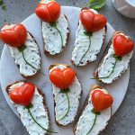 Eight bites of bread with herbed cream cheese are topped with heart-shaped tomato halves and chive stems, arranged on a round marble platter to resemble flowers—perfect for health-conscious food recipes.