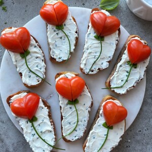Eight bites of bread with herbed cream cheese are topped with heart-shaped tomato halves and chive stems, arranged on a round marble platter to resemble flowers—perfect for health-conscious food recipes.
