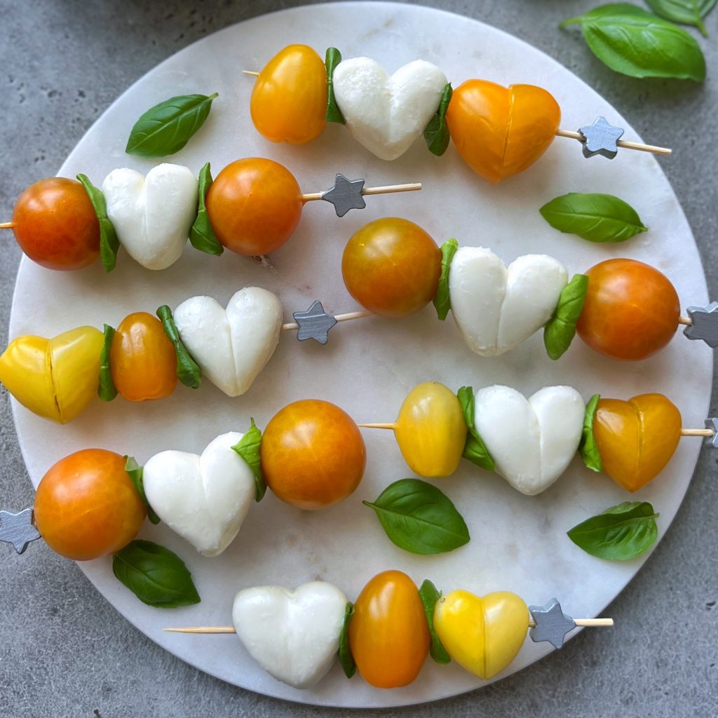 Skewers with yellow and orange cherry tomatoes, heart-shaped mozzarella, and fresh basil leaves arranged on a round marble platter. These bites make charming additions to health-focused dishes or creative food recipes. Star-shaped ends add a fun touch.