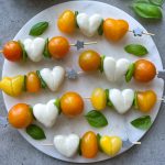 Skewers with yellow and orange cherry tomatoes, heart-shaped mozzarella, and fresh basil leaves arranged on a round marble platter. These bites make charming additions to health-focused dishes or creative food recipes. Star-shaped ends add a fun touch.