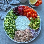 A vibrant tuna cottage cheese salad plate features diced cucumber, black olives, pickled red onions, cherry tomatoes, avocado, chopped red onion, shredded tuna, and a cottage cheese center. A towel and bowl of tomatoes and lemon sit nearby.