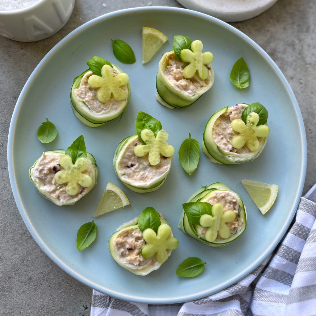 A blue plate with cucumber tuna rolls filled with a creamy mixture, garnished with small flower-shaped pieces of cucumber, fresh basil leaves, and lemon wedges on the side. A striped cloth lies nearby.