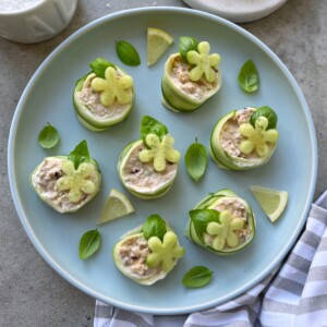 A blue plate with cucumber tuna rolls filled with a creamy mixture, garnished with small flower-shaped pieces of cucumber, fresh basil leaves, and lemon wedges on the side. A striped cloth lies nearby.
