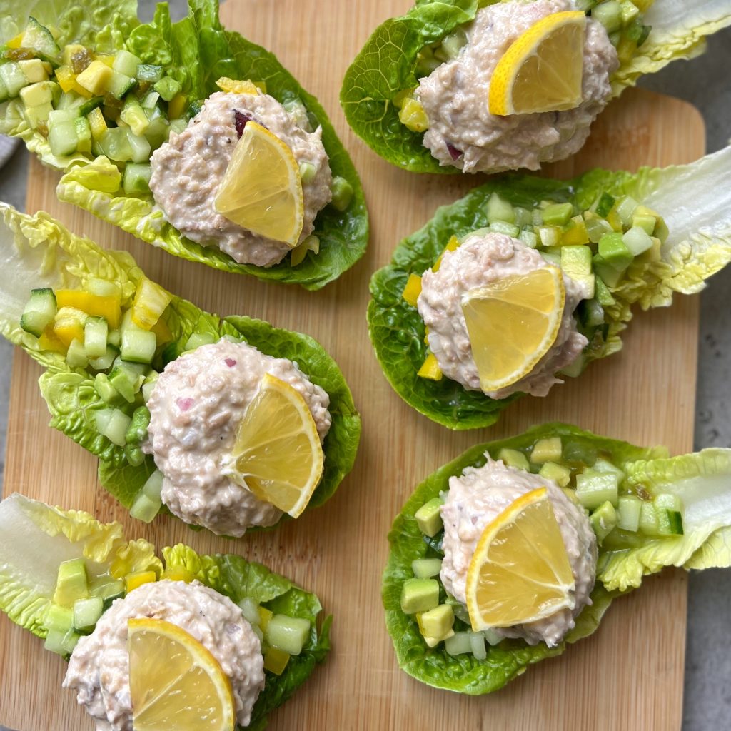 Six tuna salad lettuce boats, topped with chopped cucumber and a lemon wedge, are arranged neatly on a wooden cutting board.