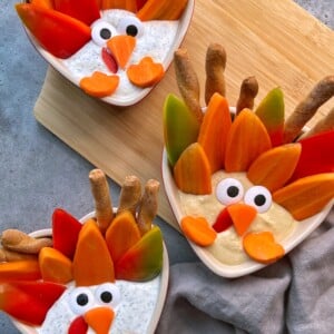 Three small bowls of healthy vegetable dip are decorated with colorful veggies, breadsticks, and cut vegetables shaped like turkeys. Displayed on a wooden board atop a gray surface.