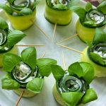 Cucumber rolls filled with cream cheese and herbs, topped with fresh basil leaves, make healthy bites arranged in a circle on a plate with toothpicks holding each roll together. Perfect for those who love quick food recipes.
