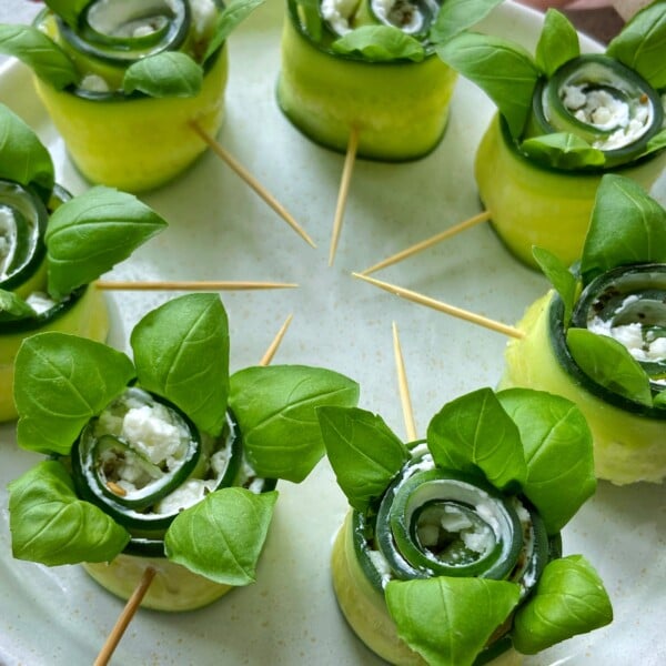 Cucumber rolls filled with cream cheese and herbs, topped with fresh basil leaves, make healthy bites arranged in a circle on a plate with toothpicks holding each roll together. Perfect for those who love quick food recipes.