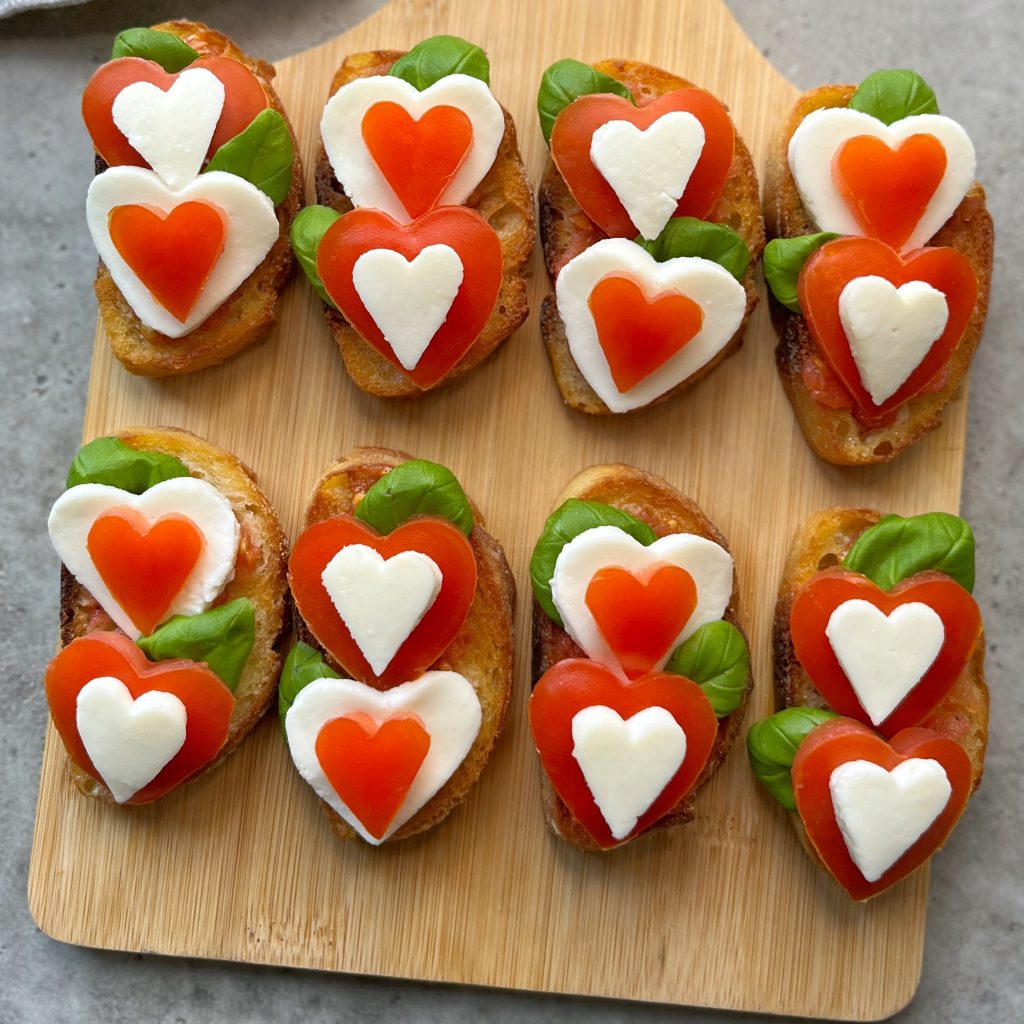 Eight bruschetta bites topped with basil, mozzarella, and heart-shaped tomato slices are arranged neatly on a wooden board. The toppings form a double heart pattern on each piece—perfect for sharing health and love through beautiful dishes.