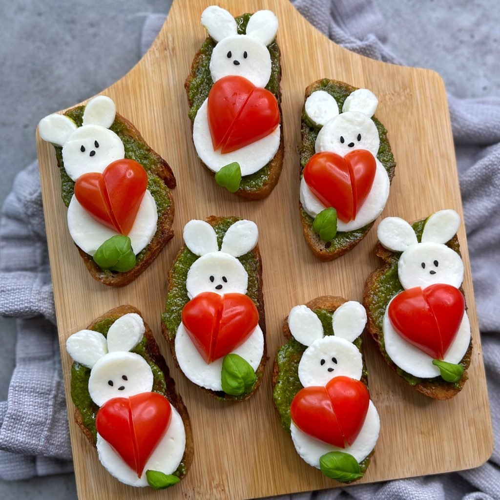 Bruschetta slices topped with mozzarella cheese, cherry tomatoes shaped like bunnies holding red hearts, basil leaves, and pesto create adorable bites arranged on a wooden board—perfect for adding fun to your favorite food recipes.