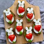 Bruschetta slices topped with mozzarella cheese, cherry tomatoes shaped like bunnies holding red hearts, basil leaves, and pesto create adorable bites arranged on a wooden board—perfect for adding fun to your favorite food recipes.