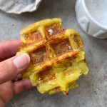A hand holding a crispy, golden-brown waffle-shaped potato bite with flakes of sea salt on top; a white bowl is visible in the background on a gray surface—perfect inspiration for new food recipes.