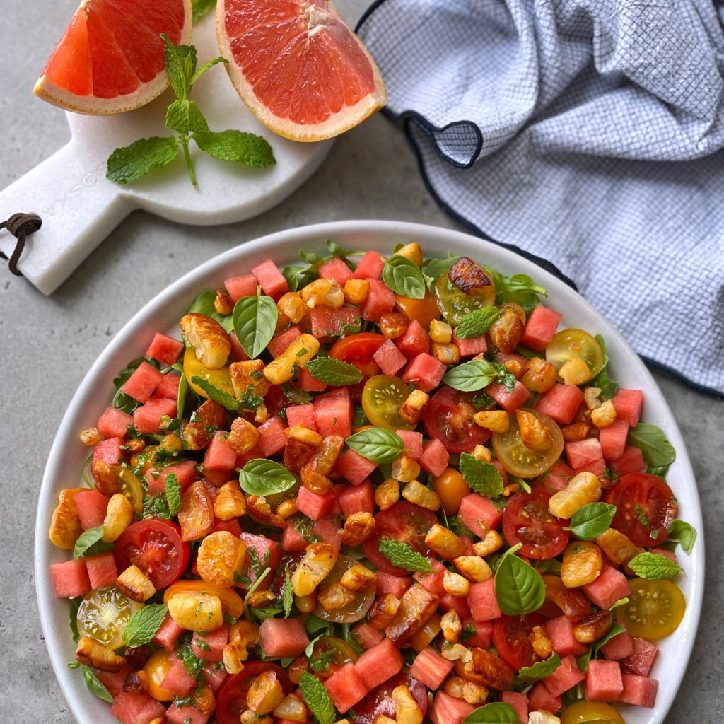 A bowl of colorful salad with watermelon cubes, cherry tomatoes, orange segments, greens, and fresh herbs—perfect for health-focused food recipes. Nearby are grapefruit slices on a board and a blue checkered cloth.