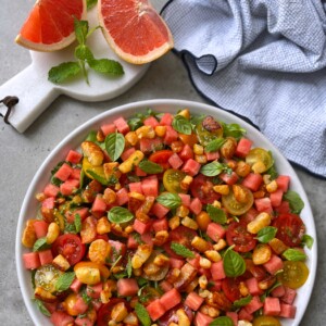 A bowl of colorful salad with watermelon cubes, cherry tomatoes, orange segments, greens, and fresh herbs—perfect for health-focused food recipes. Nearby are grapefruit slices on a board and a blue checkered cloth.