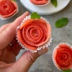 A hand holds a cupcake liner with thin slices of watermelon arranged in a rose shape, garnished with a small mint leaf. These refreshing bites make for beautiful, health-inspired dishes, with more watermelon roses displayed on the gray surface behind.