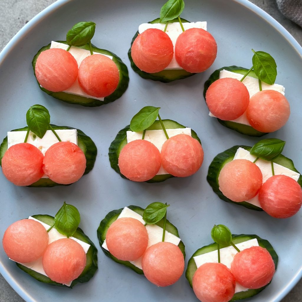 Slices of cucumber topped with white cheese, two small round pink watermelon bites, and a fresh basil leaf are arranged neatly on a gray plate—perfect for health-conscious food lovers seeking refreshing recipes.
