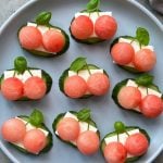 A plate of healthy appetizers with cucumber slices topped with white cheese, round watermelon balls, and a basil leaf on each—perfect for refreshing bites arranged neatly in rows.