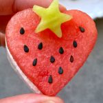 A hand holds a heart-shaped slice of watermelon feta bite, topped with a small, yellow star-shaped fruit and decorated with black seeds.
