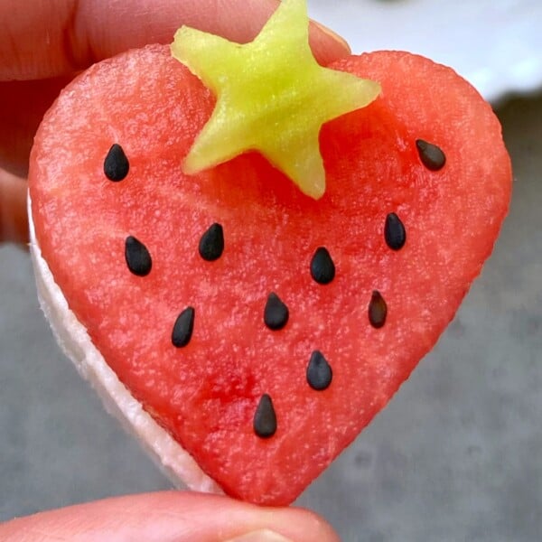 A hand holds a heart-shaped slice of watermelon feta bite, topped with a small, yellow star-shaped fruit and decorated with black seeds.