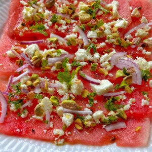 Thin slices of watermelon feta carpaccio are topped with crumbled feta, red onion, chopped pistachios, and fresh mint, all beautifully arranged on a white plate.