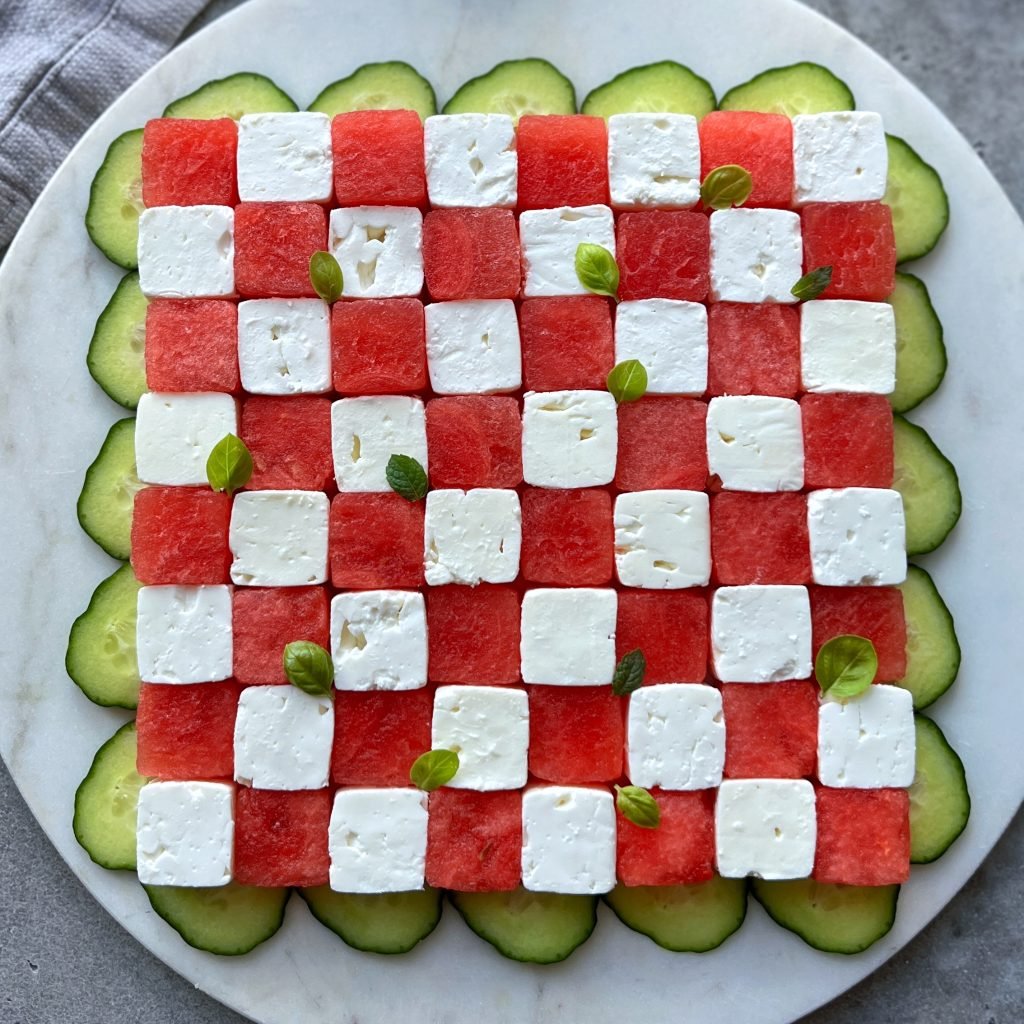 A square salad arranged like a checkerboard with alternating cubes of watermelon and feta, garnished with small basil leaves, and surrounded by overlapping cucumber slices on a round white plate—perfect for fresh, healthy bites.
