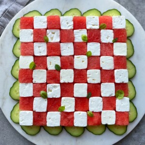 A square salad arranged like a checkerboard with alternating cubes of watermelon and feta, garnished with small basil leaves, and surrounded by overlapping cucumber slices on a round white plate—perfect for fresh, healthy bites.