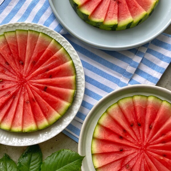 Three plates with neatly sliced watermelon circles are placed on a blue and white striped cloth. These refreshing bites make for vibrant, healthy dishes, with green rind visible and a few seeds and leaves nearby.