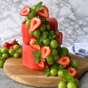 Two stacked watermelon fruit cake blocks are decorated with green grapes, halved strawberries, and mint leaves, arranged on a wooden board with a gray marble background.