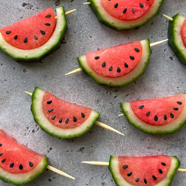 Slices of watermelon with green rind and black seeds are arranged on a gray surface; some pieces are skewered with wooden sticks and spaced evenly in a pattern, making for refreshing bites perfect for healthy dishes or summer food recipes.