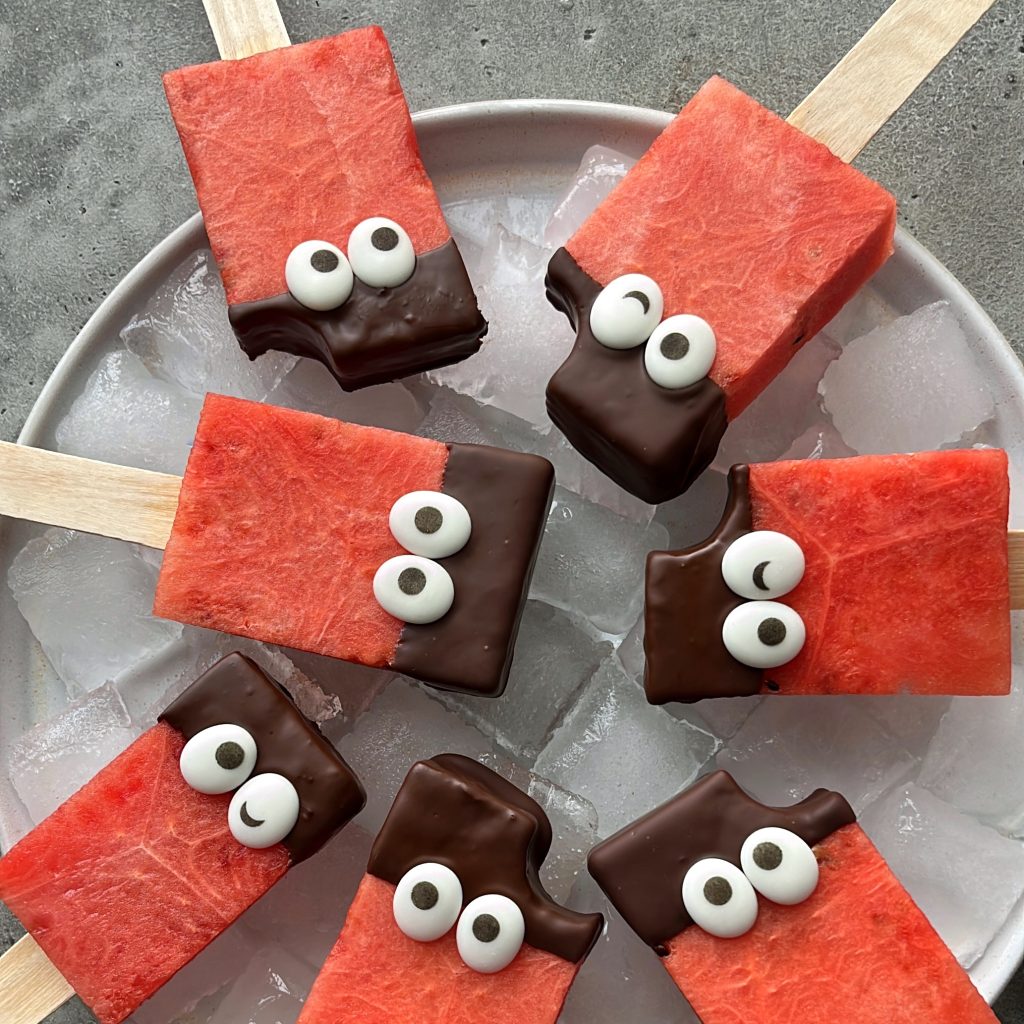 Watermelon popsicles with chocolate-dipped tops and candy eyes are arranged in a circle on a plate of ice, creating playful and whimsical bites that add fun to your food recipes.