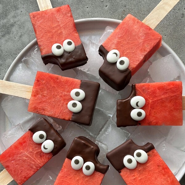 Rectangular watermelon popsicles on sticks, partially dipped in chocolate and decorated with candy eyes, are arranged on a plate of ice. One popsicle has a small bite taken out—making these fun bites perfect for healthy food recipes.