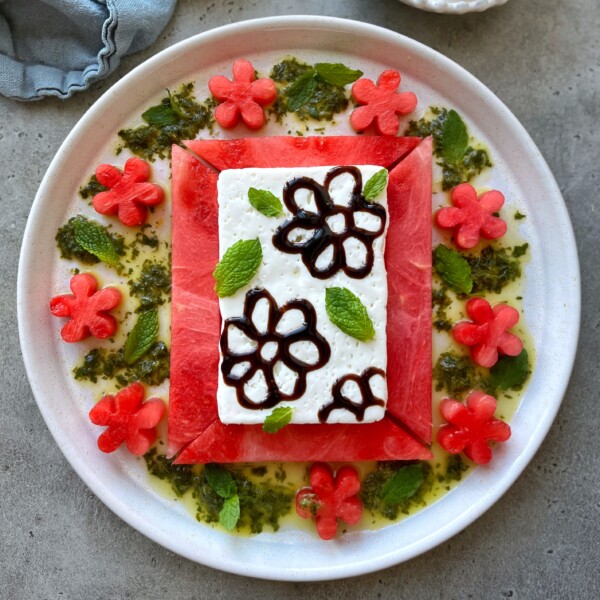 A white plate with a rectangular slice of watermelon topped with feta cheese, decorated with chocolate flower designs, surrounded by flower-shaped watermelon bites, mint leaves, and green sauce—perfect for creative food recipes and fresh summer dishes.
