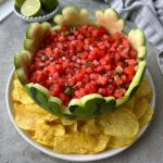 A hollowed-out watermelon bowl filled with fresh watermelon salsa sits on a white plate surrounded by yellow tortilla chips. Lime wedges and a gray cloth are nearby.