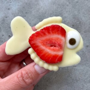 A hand holds a fish-shaped treat made of white chocolate, with a strawberry slice as the fish’s body and a small candy eye, offering creative bites for those who love playful food recipes against a gray background.