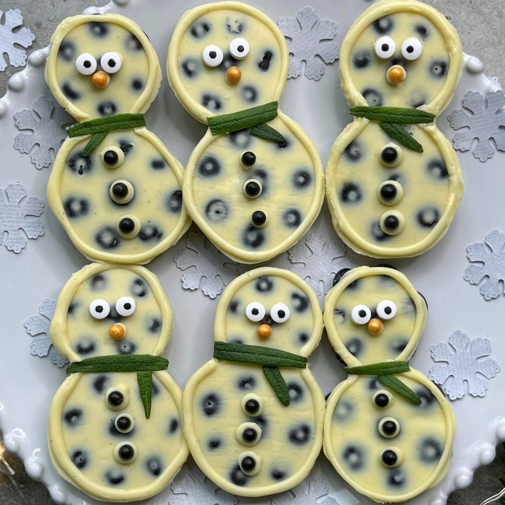 Six snowman treats made with white chocolate and blueberries, decorated with candy eyes, orange sprinkle noses, black sprinkle buttons, and green rosemary scarves.