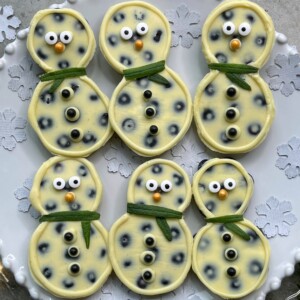 Six snowman-shaped bites with white chocolate and dark spots, decorated with candy eyes, orange noses, black buttons, and green candy scarves, are arranged on a decorative white plate with snowflake patterns—perfect for festive food recipes.