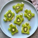 Six turtle-shaped bites on a blue plate, crafted from slices of kiwi for shells and green grapes for heads and legs, with candy eyes on top—an adorable addition to your collection of fun food recipes and healthy snacks.