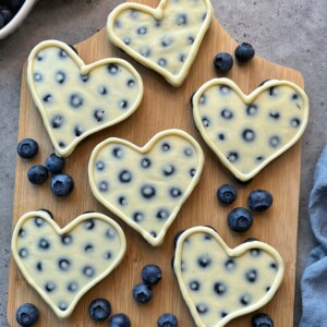 Six heart-shaped cheesecakes with embedded blueberries are arranged on a wooden board, creating delicious bites surrounded by fresh blueberries on a gray surface. Perfect for anyone who loves health-conscious food recipes.