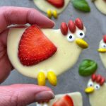 A hand holds a white chocolate treat shaped like a chicken, decorated with a strawberry slice as a wing, yellow candies as feet and beak, red candies as a comb, and candy eyes—perfect for creative food recipes or festive bites. Basil leaves and treats are in the background.