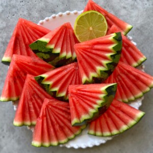 A white plate holds neatly arranged, fan-cut watermelon slices with green rinds and a halved lime on top, all displayed on a gray surface—perfect for healthy bites or as inspiration for fresh food recipes.