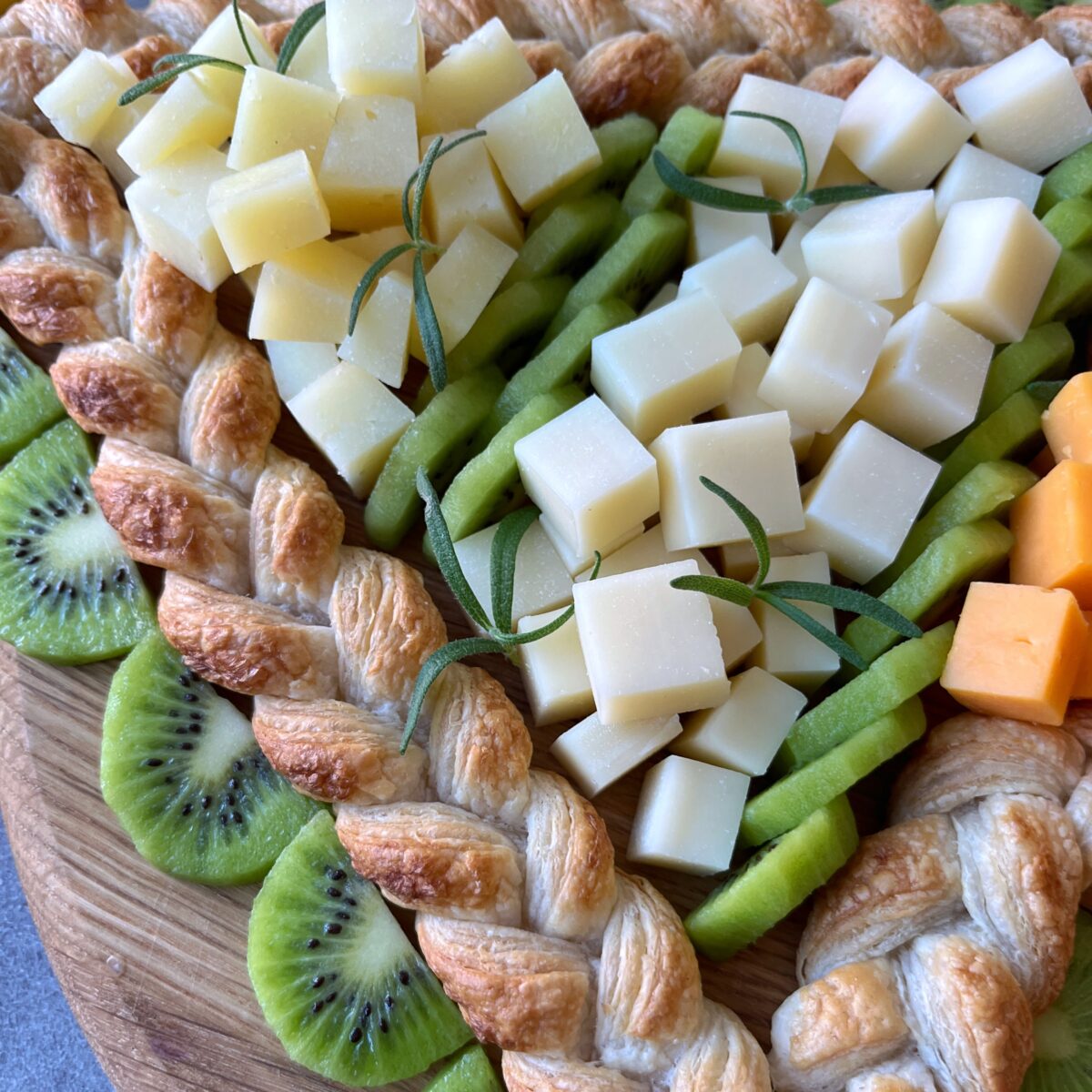 A wooden board with braided puff pastry, sliced kiwi, and cubes of various cheeses, garnished with sprigs of rosemary.