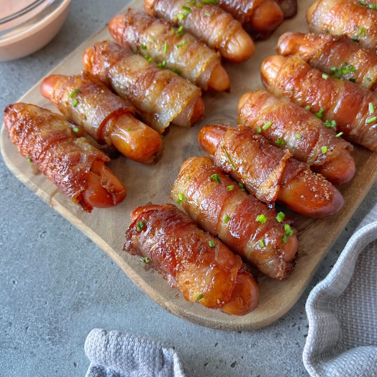 Mini sausages wrapped in crispy bacon are arranged on a wooden board, garnished with chopped herbs, next to a dipping sauce.