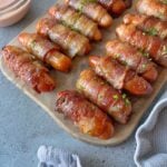 Mini sausages wrapped in crispy bacon are arranged on a wooden board, garnished with chopped herbs, next to a dipping sauce.