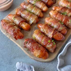 Mini sausages wrapped in crispy bacon are arranged on a wooden board, garnished with chopped herbs, next to a dipping sauce.
