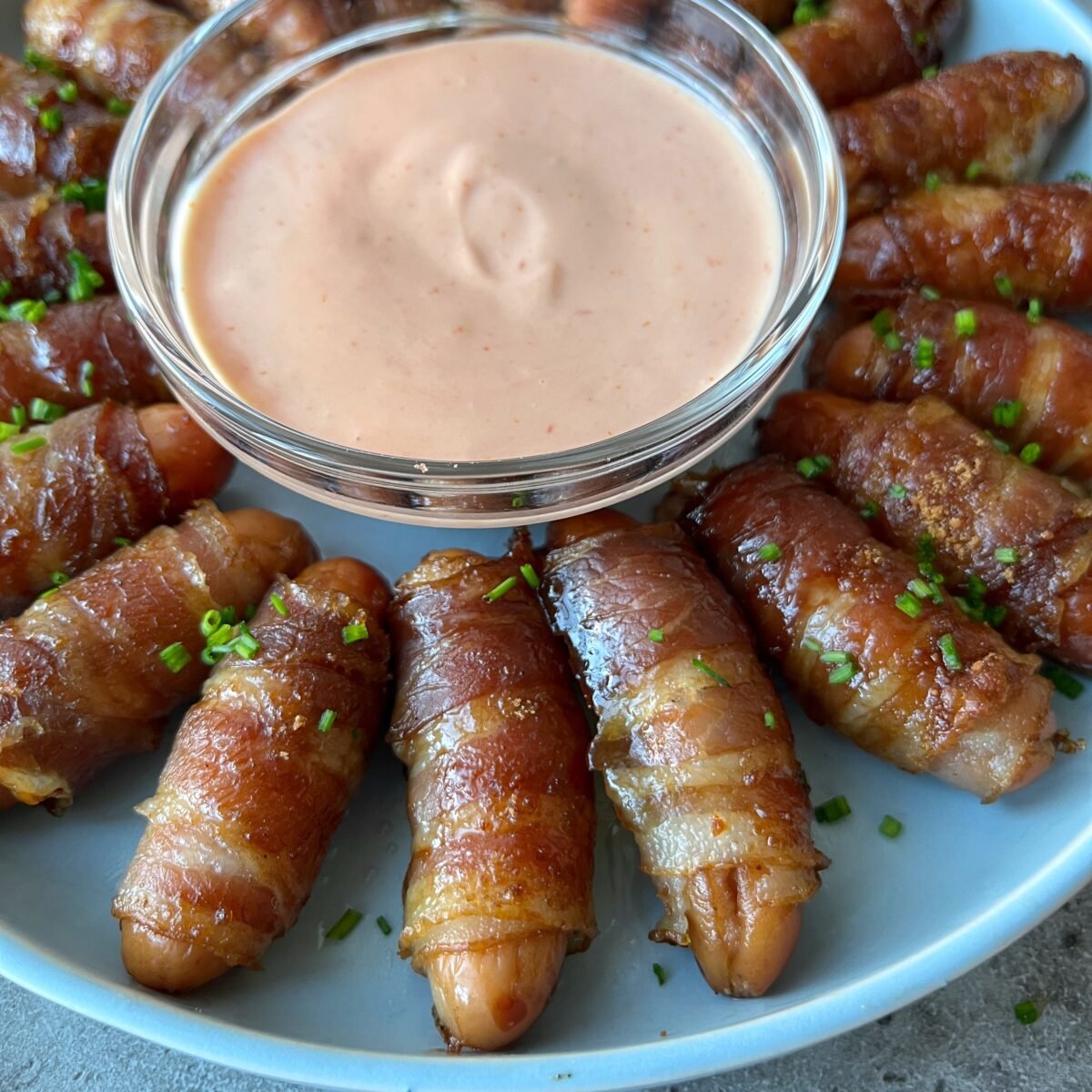 A plate of bacon-wrapped mini sausages arranged in a circle around a bowl of creamy pink dipping sauce, garnished with chopped chives.